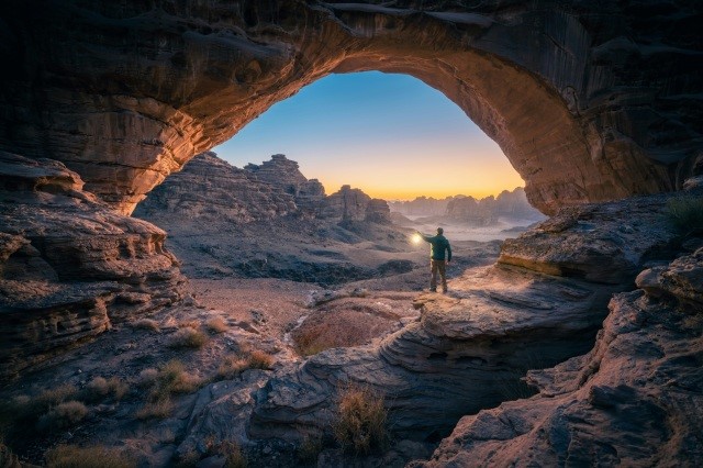 Man holding up a lantern at the mouth of a cave, surveying a canyon below.