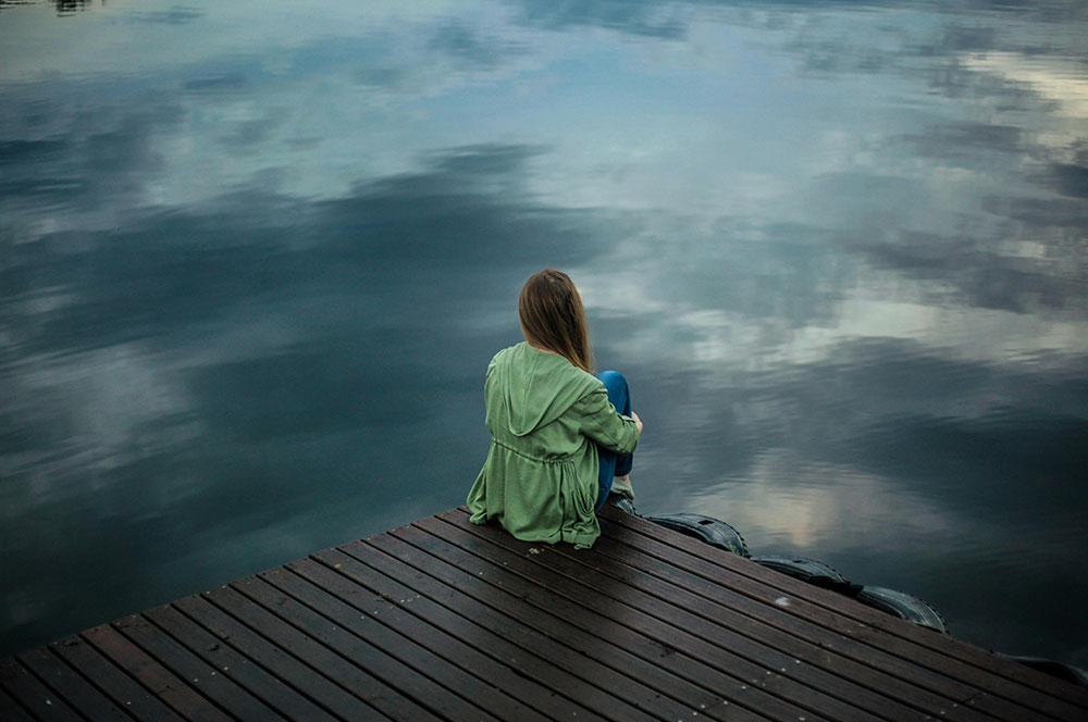 Lady sitting on a dock, looking out on a small lake, with her arm wrapped around her knees.